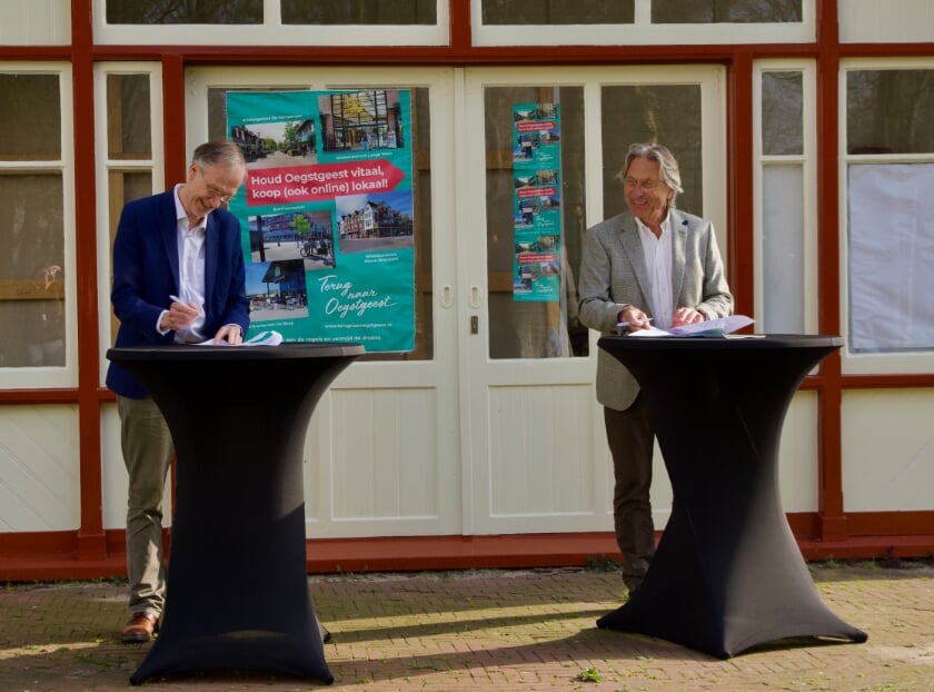 <p>Wethouder Economische Zaken Jan Nieuwenhuis (links) en de voorzitter van Dorpsmarketing Oegstgeest, Hans Ludo van Mierlo, ondertekenen de nieuwe overeenkomst bij het gemeentehuis Oegstgeest.</p>